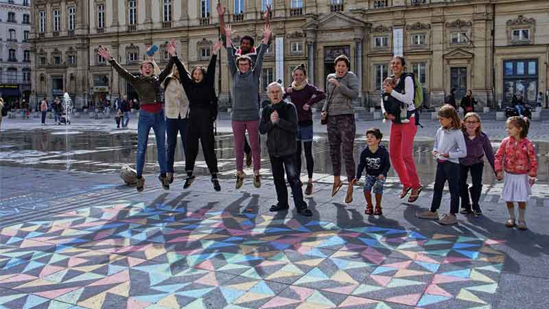 Un groupe d'adultes et d'enfants se tient sur une place de la ville devant un pavage coloré dessiné à la craie sur le sol.