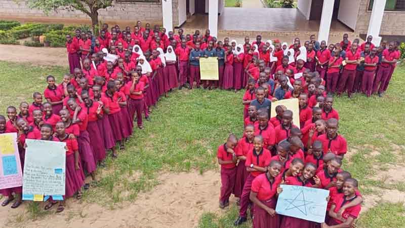 A large group of young students in red school uniforms standing in front of the school building, forming a large letter Pi.